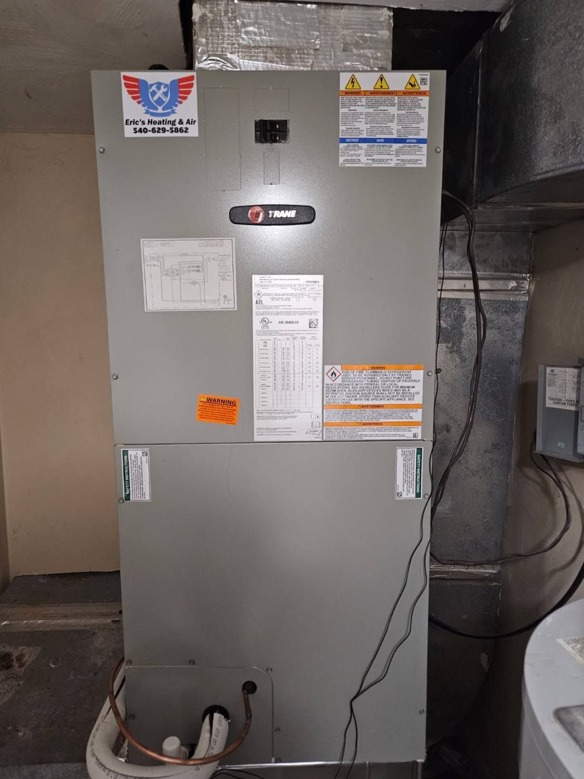 Gray HVAC furnace in a utility room with labels and ductwork, near a water heater and floor pipes
