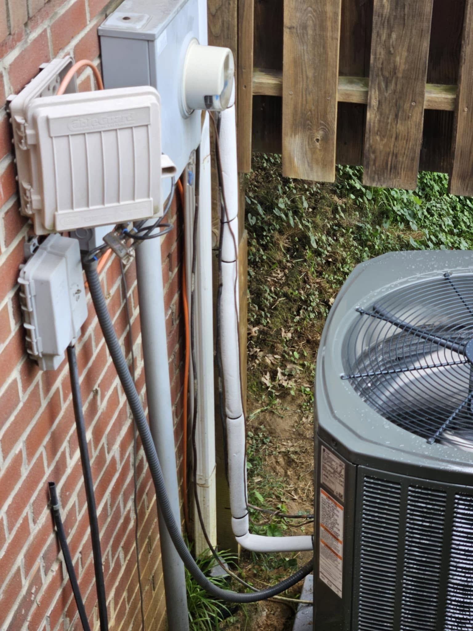 Outdoor air conditioner beside a brick wall, with white utility pipes and a fenced yard nearby