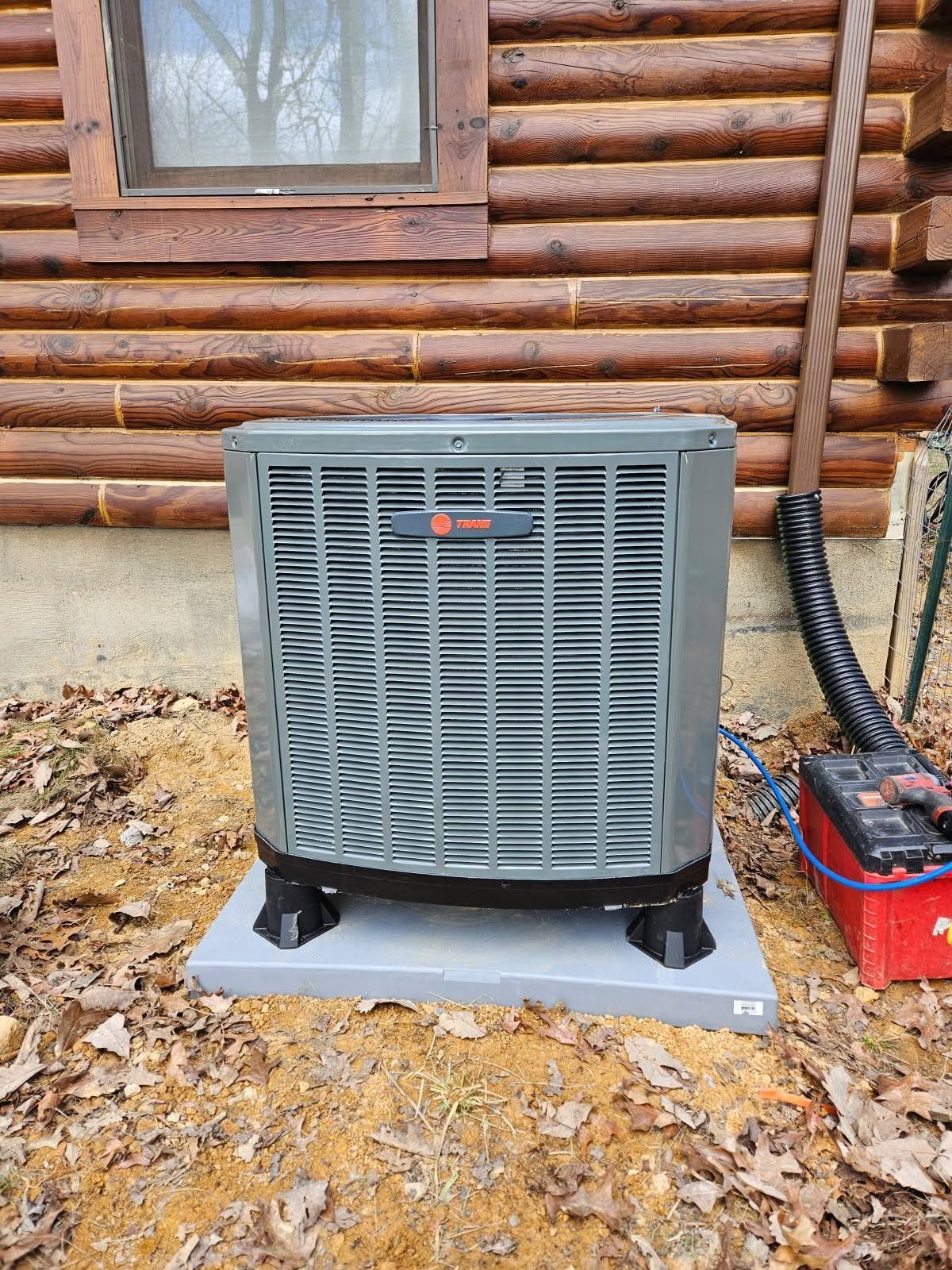 Outdoor HVAC condenser unit beside a log cabin on a concrete pad with leaves on the ground