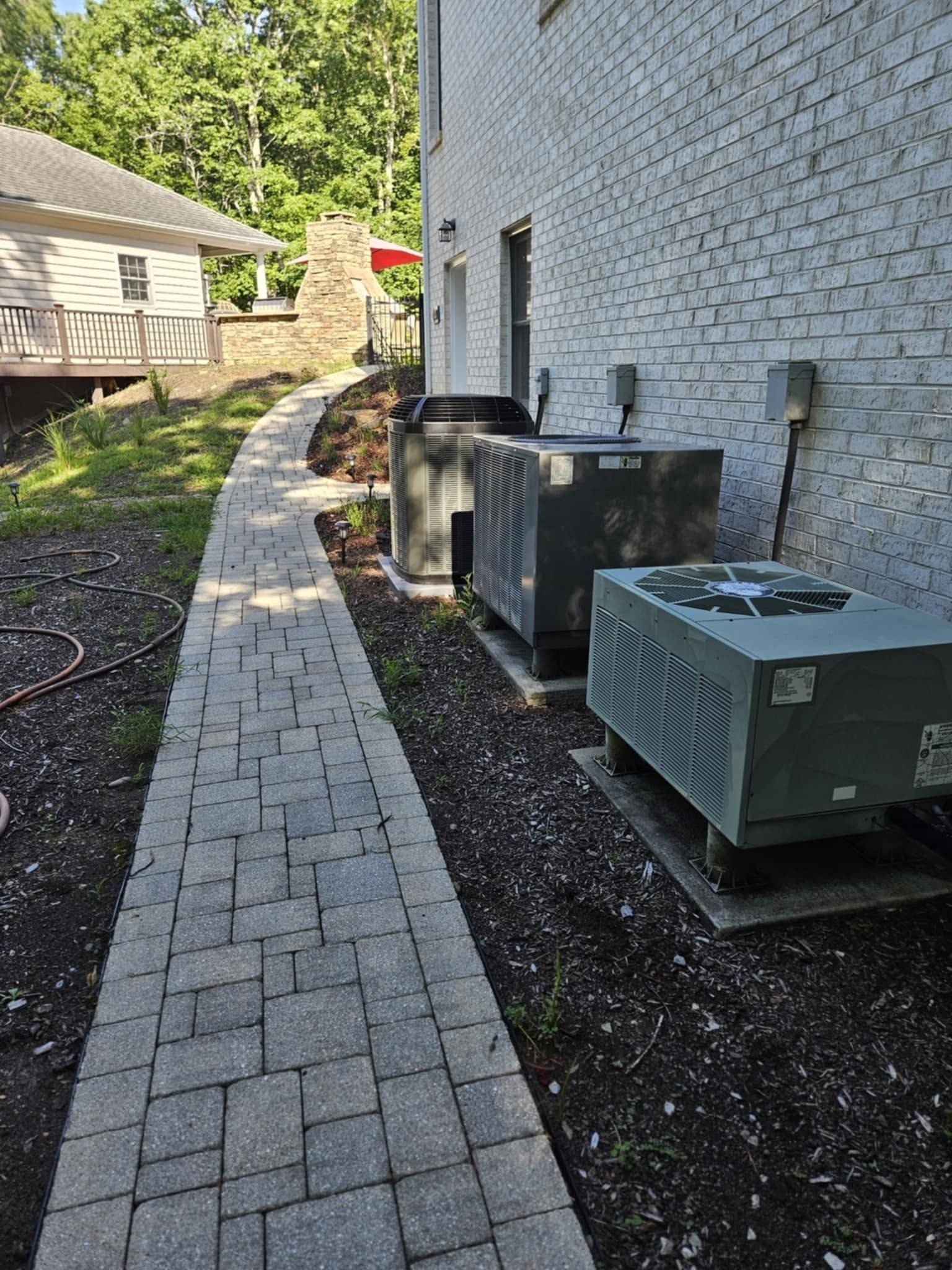 Curved brick walkway beside a house, with outdoor HVAC units along the wall and landscaping on the side.