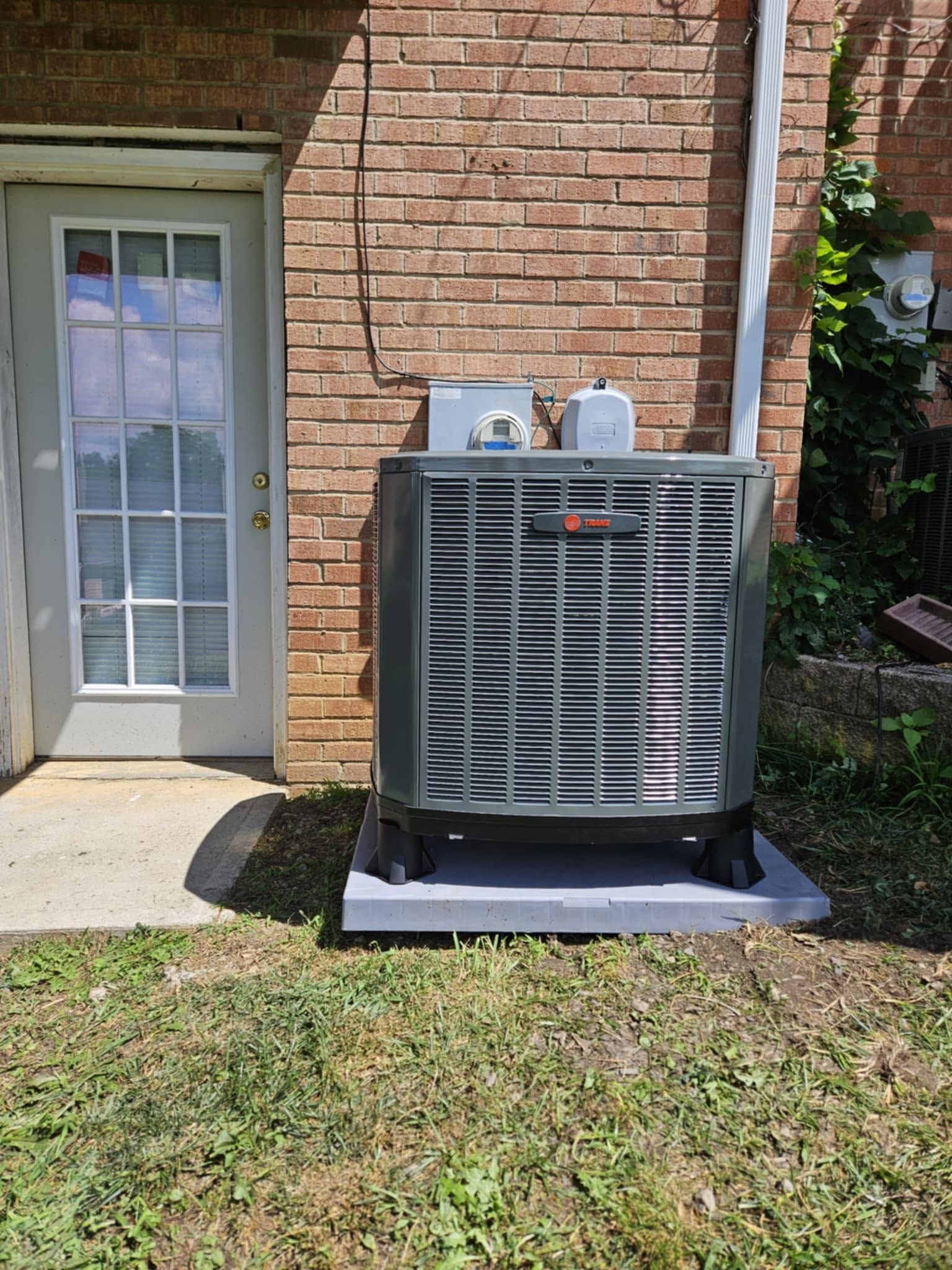Outdoor HVAC unit beside a brick house wall and glass door, set on a concrete pad in a grassy yard