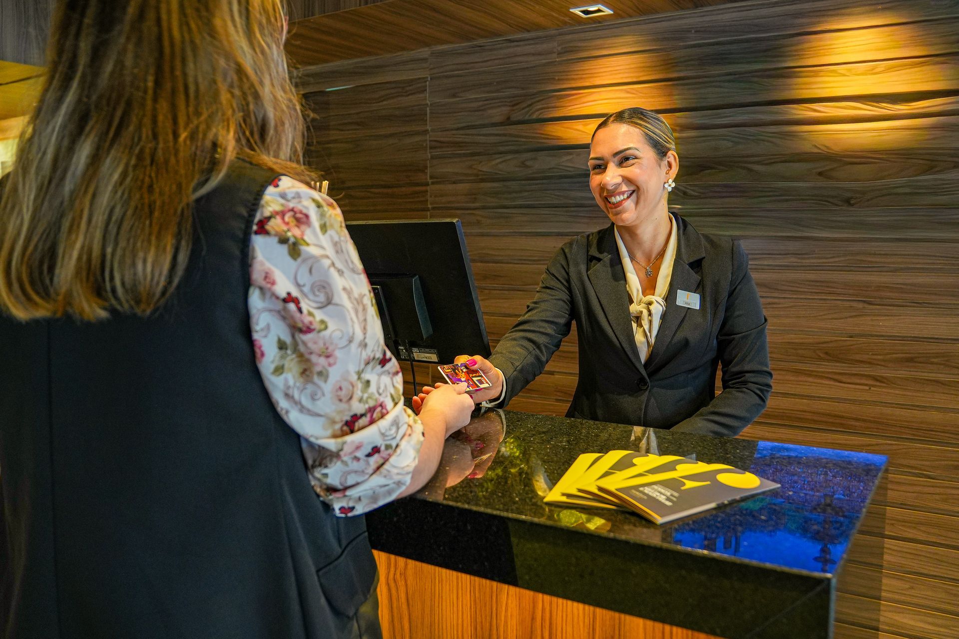 Uma recepcionista de hotel sorri enquanto entrega um cartão-chave a um hóspede em um balcão de recepção com painéis de madeira.