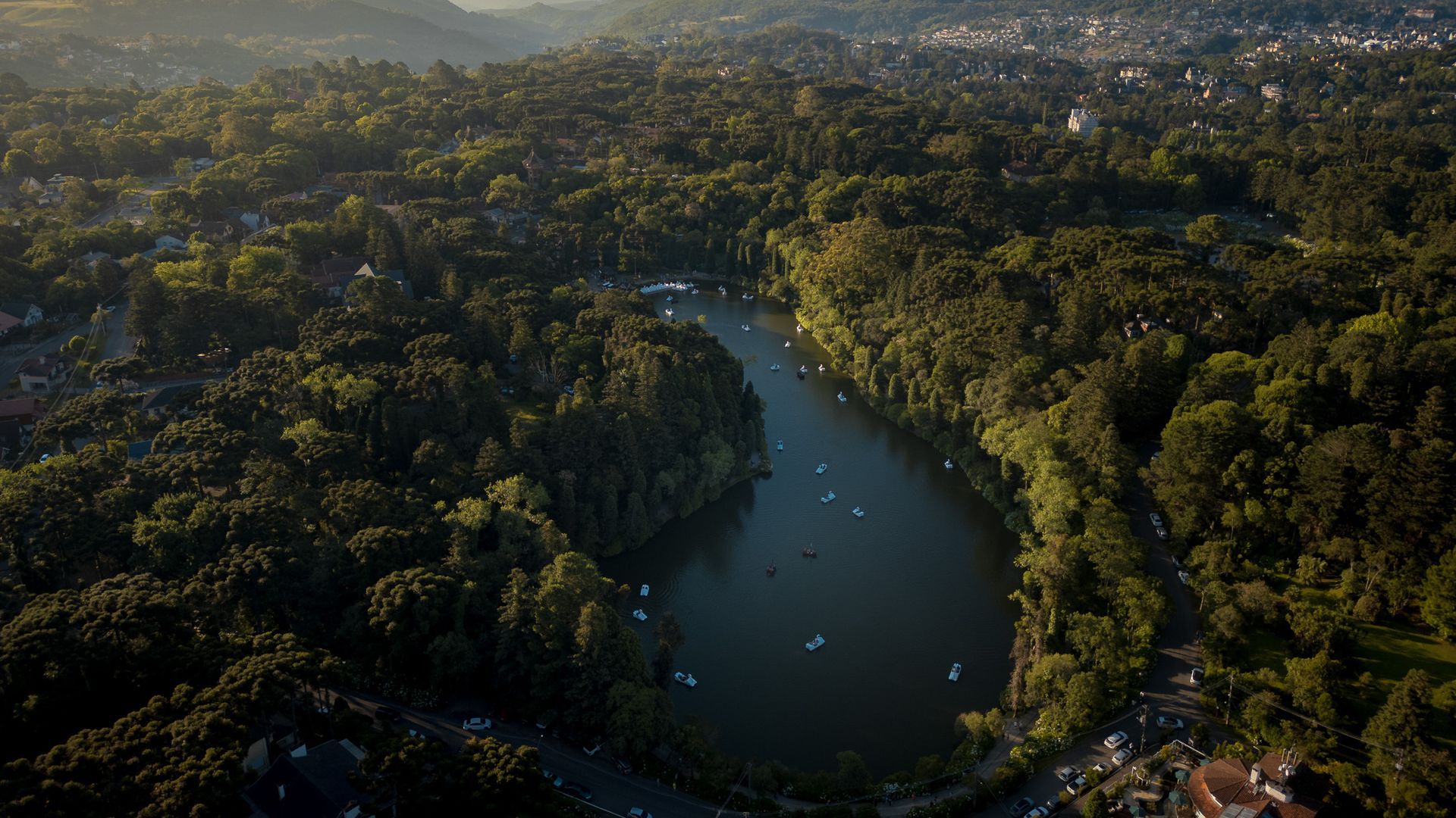 Vista aérea de um lago escuro e alongado, cercado por uma densa floresta verde ao pôr do sol, com pequenos barcos flutuando na água.