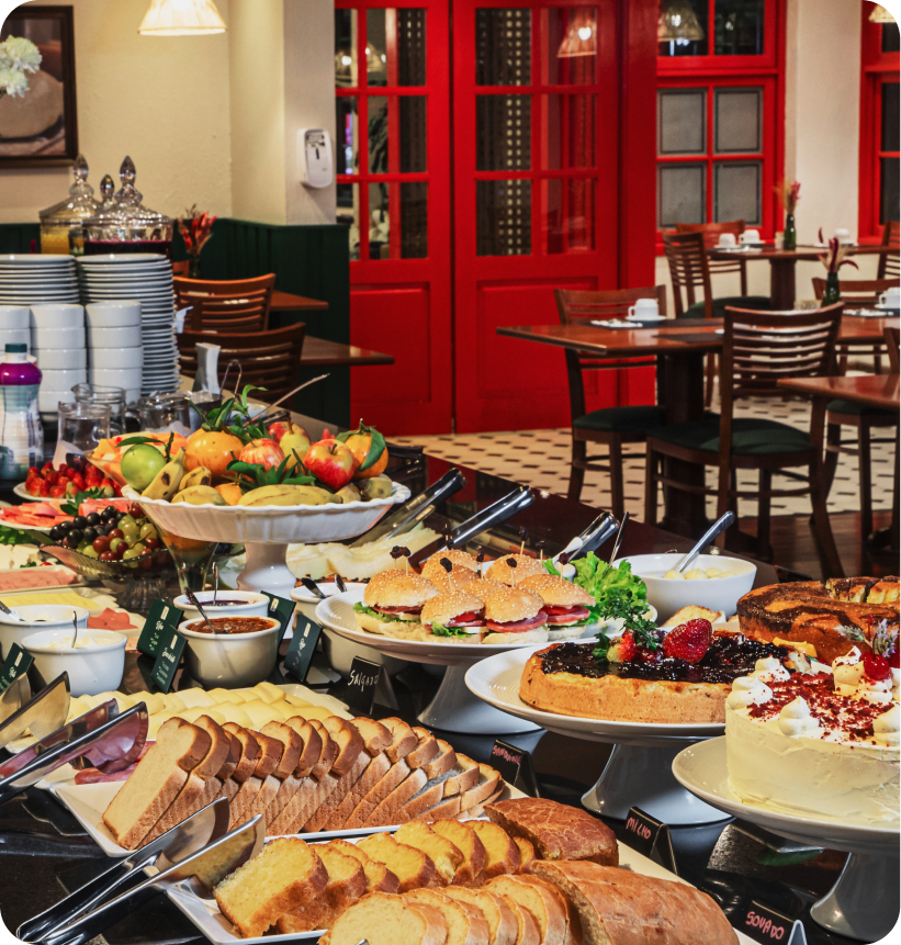 Mesa de buffet com doces, frutas e pratos em um café iluminado com portas vermelhas e mesas de jantar vazias.