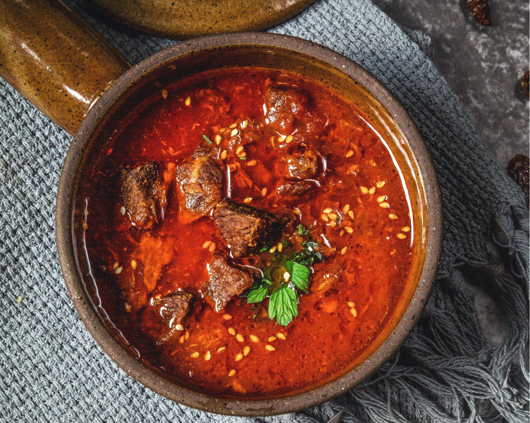 Tigela de ensopado vermelho com pedaços de carne e ervas sobre uma toalha de mesa rústica.