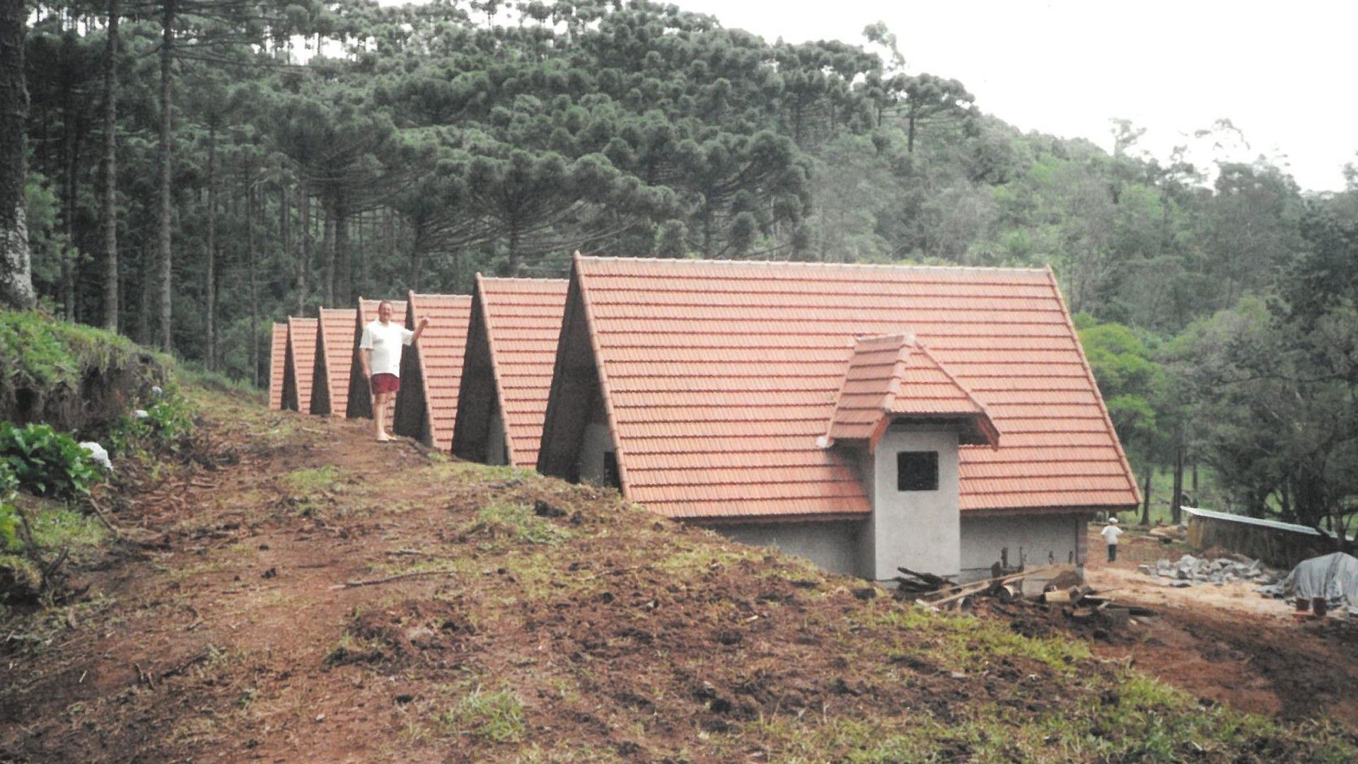 Uma fileira de cabanas idênticas com telhados de telha vermelha situadas em uma encosta arborizada, com uma pessoa em pé perto de uma delas.