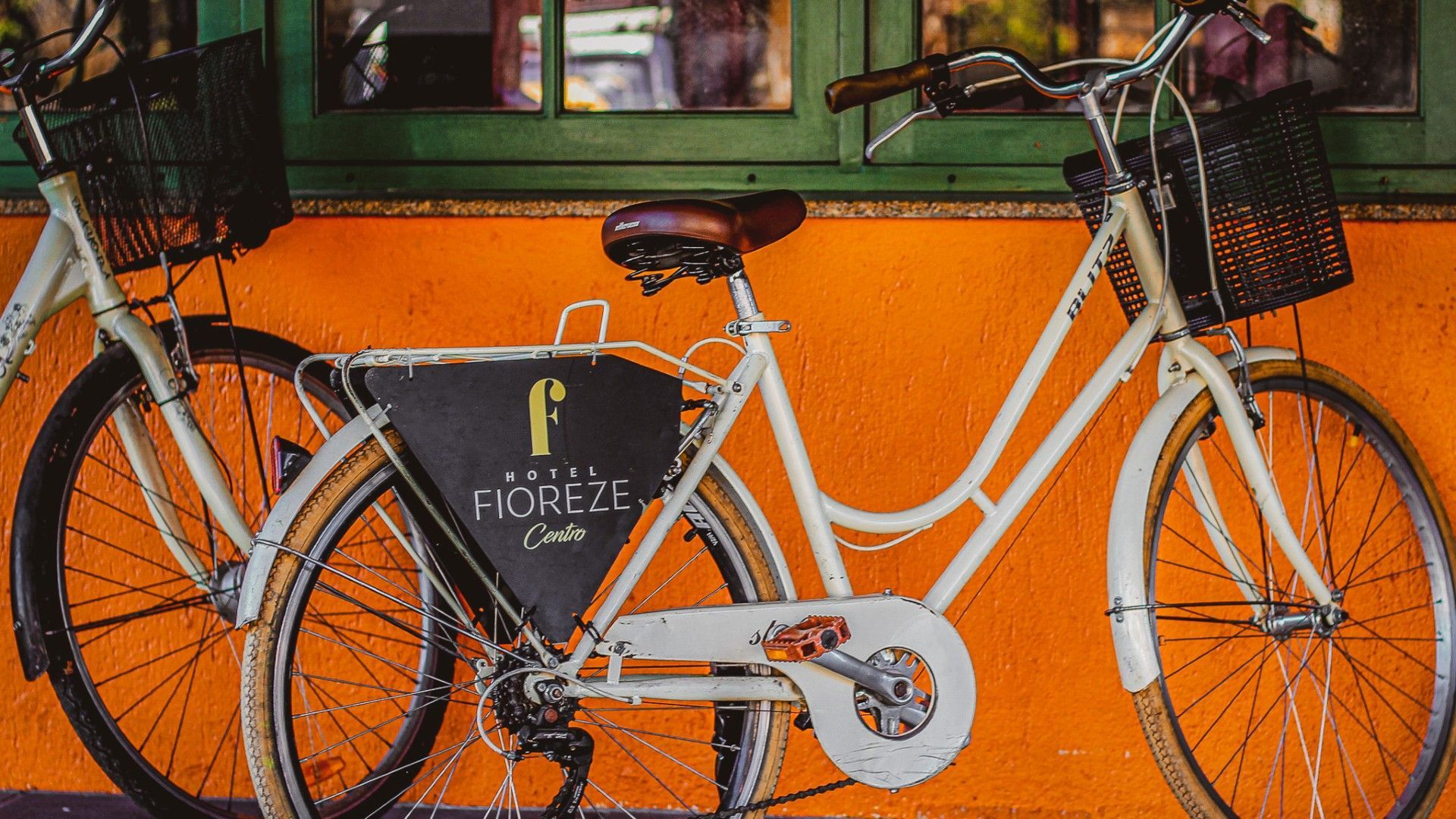 Duas bicicletas antigas cor creme com cestas dianteiras estacionadas contra uma parede laranja vibrante.