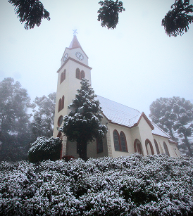 Torre da igreja e capela cobertas de neve em meio a árvores geladas sob um céu cinzento.