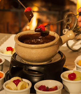 Panela de fondue de chocolate com morangos e frutas no espeto, rodeada por tigelas de guloseimas para mergulhar.