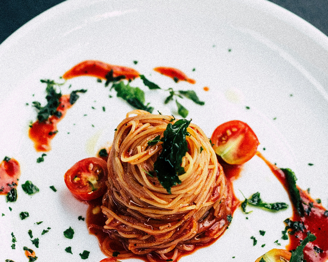 Ninho de espaguete em prato branco com molho de tomate, tomates-cereja e ervas aromáticas.