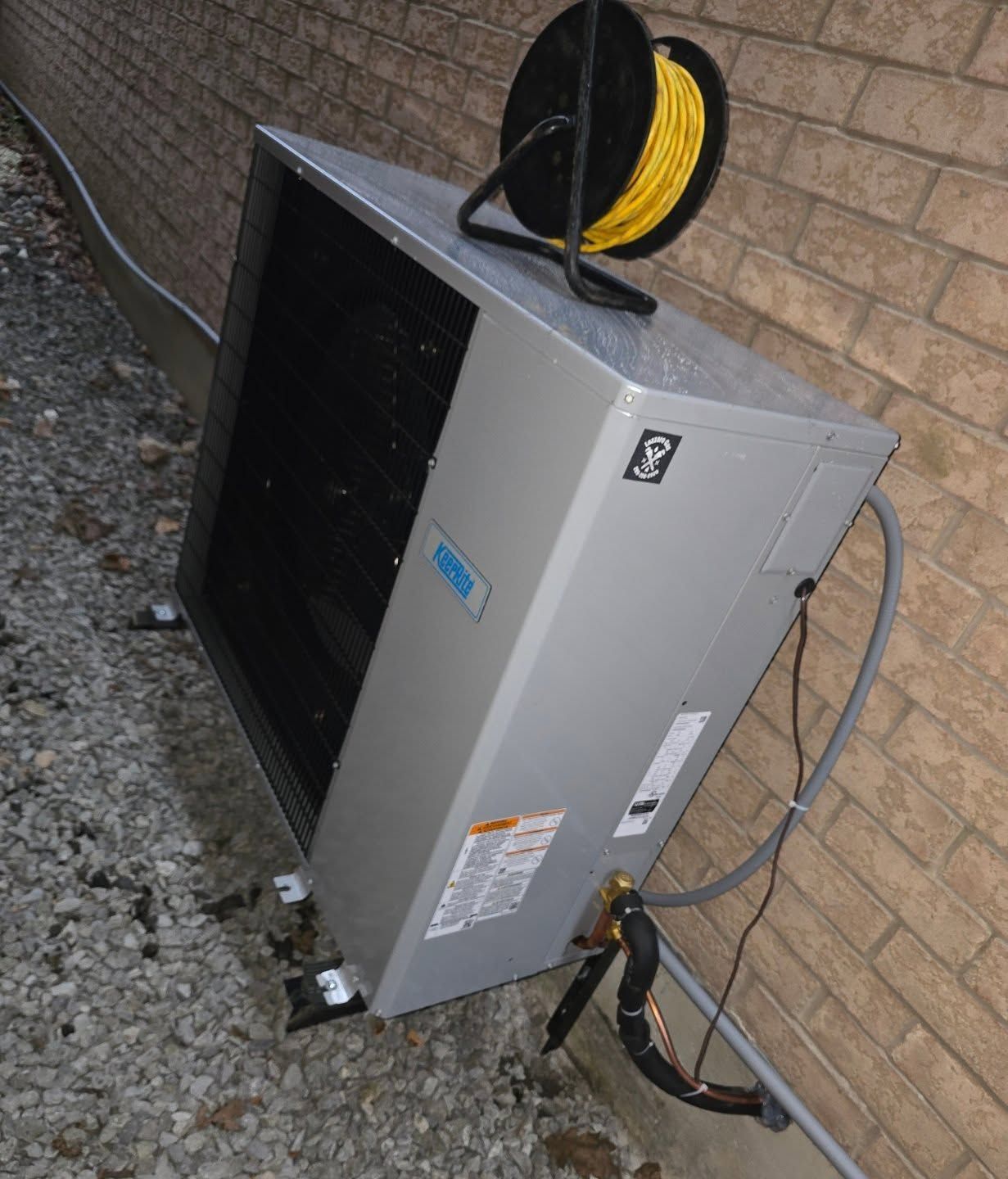 Outdoor HVAC unit against a brick wall, with a yellow extension cord spool on top.