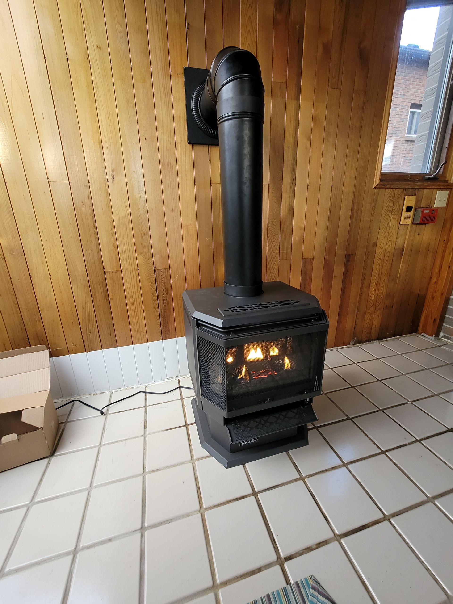 Black wood stove with lit fire and stovepipe in a tiled room with wood-paneled walls.