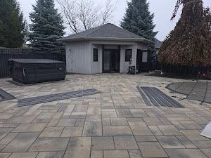 Patio with hot tub and small building with dark roof, pavers in foreground, trees in background.