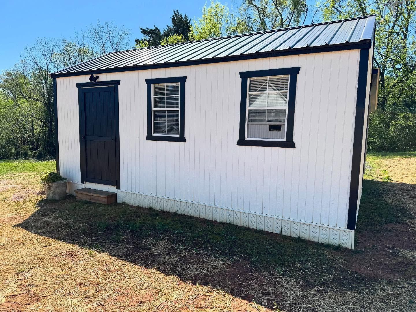 White shed with black trim, door, and metal roof. Two windows and a door on the front, set in a grassy outdoor area.