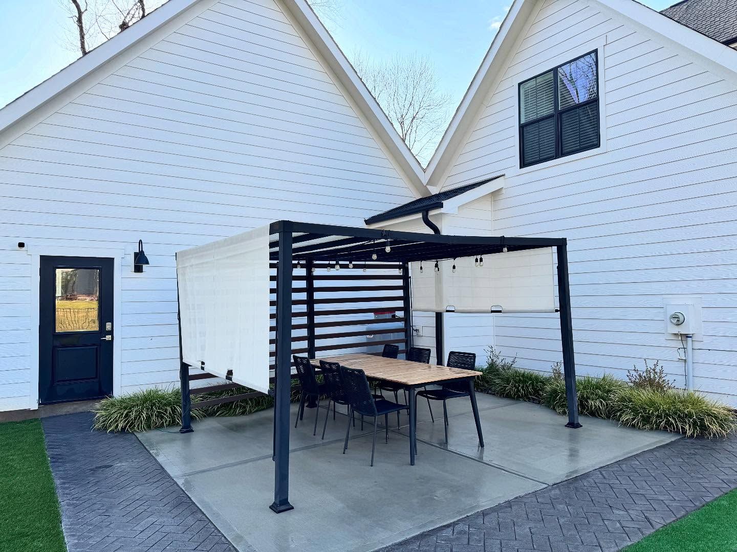 A modern outdoor dining area with a pergola, table, and chairs, set between two white buildings.