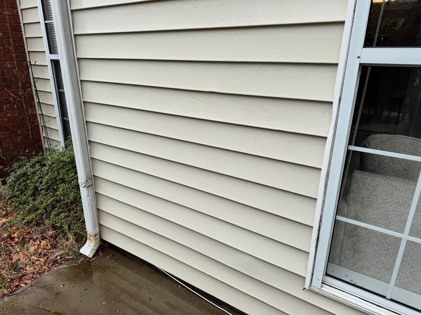 Beige siding on a building with a white window frame and a downspout. A concrete sidewalk is in the foreground.