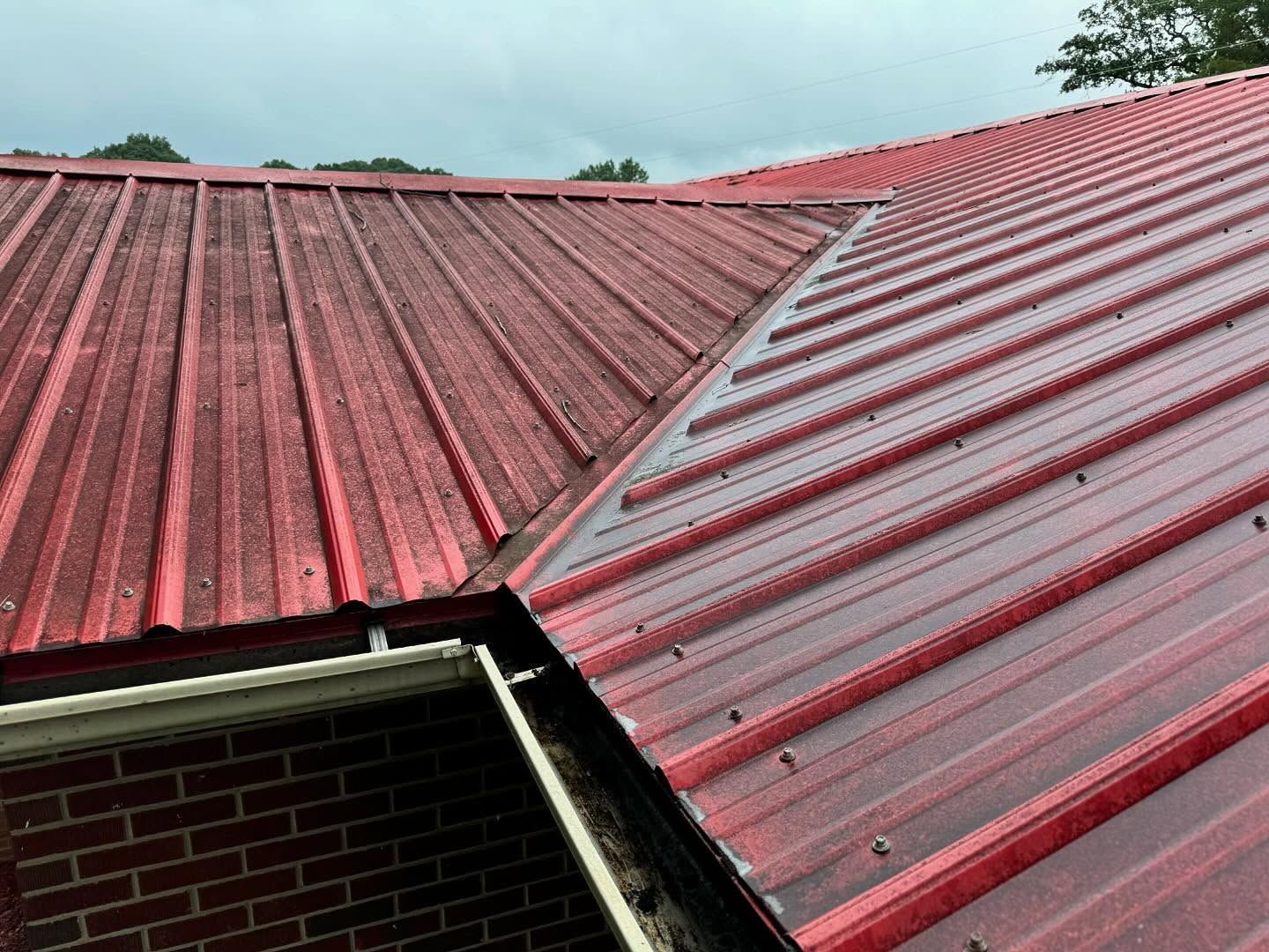 Red metal roof with visible ridges meeting at a corner, next to a brick building. Gutters are visible along the lower edge.