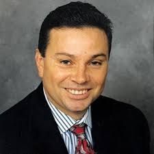 Smiling man in a dark suit, striped shirt, and red tie against a gray background