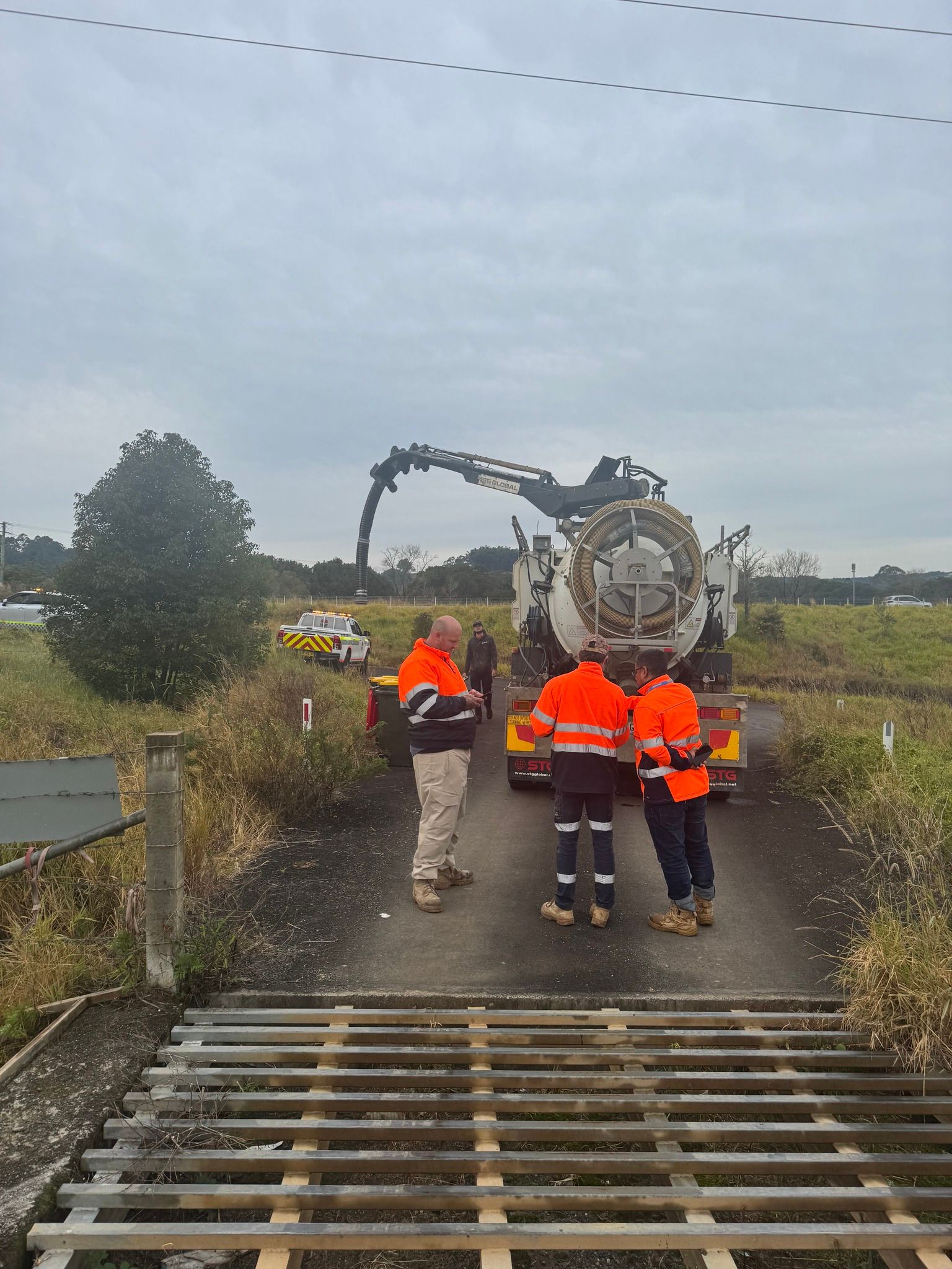 Workers in Orange Vests Observe a Tanker Truck Unloading Tar, Standing Near a Cattle Guard on a Rural Road — Tweed Heads Leak Detection in Casuarina, NSW