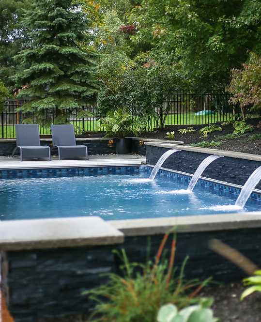 Backyard pool with two gray lounge chairs and a waterfall spilling into blue water, surrounded by greenery