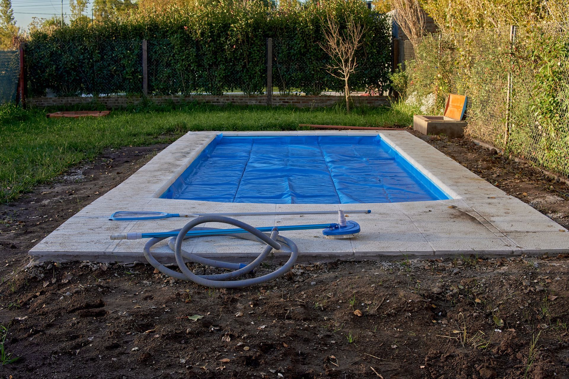 Small rectangular in-ground pool with blue liner, hose, and concrete deck in a fenced backyard