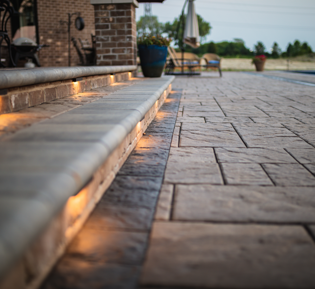 A brick walkway with lights on the steps leading to a swimming pool.