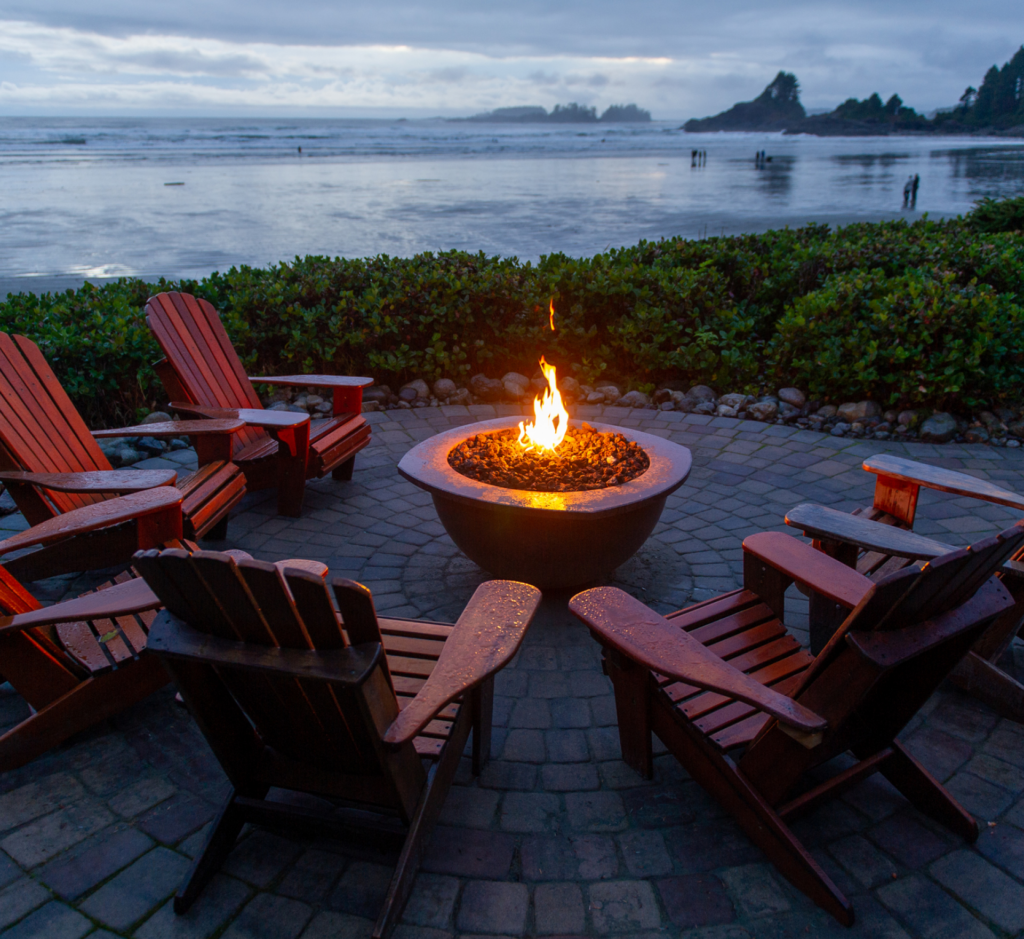 A fire pit is surrounded by wooden chairs on a patio with a view of the ocean.