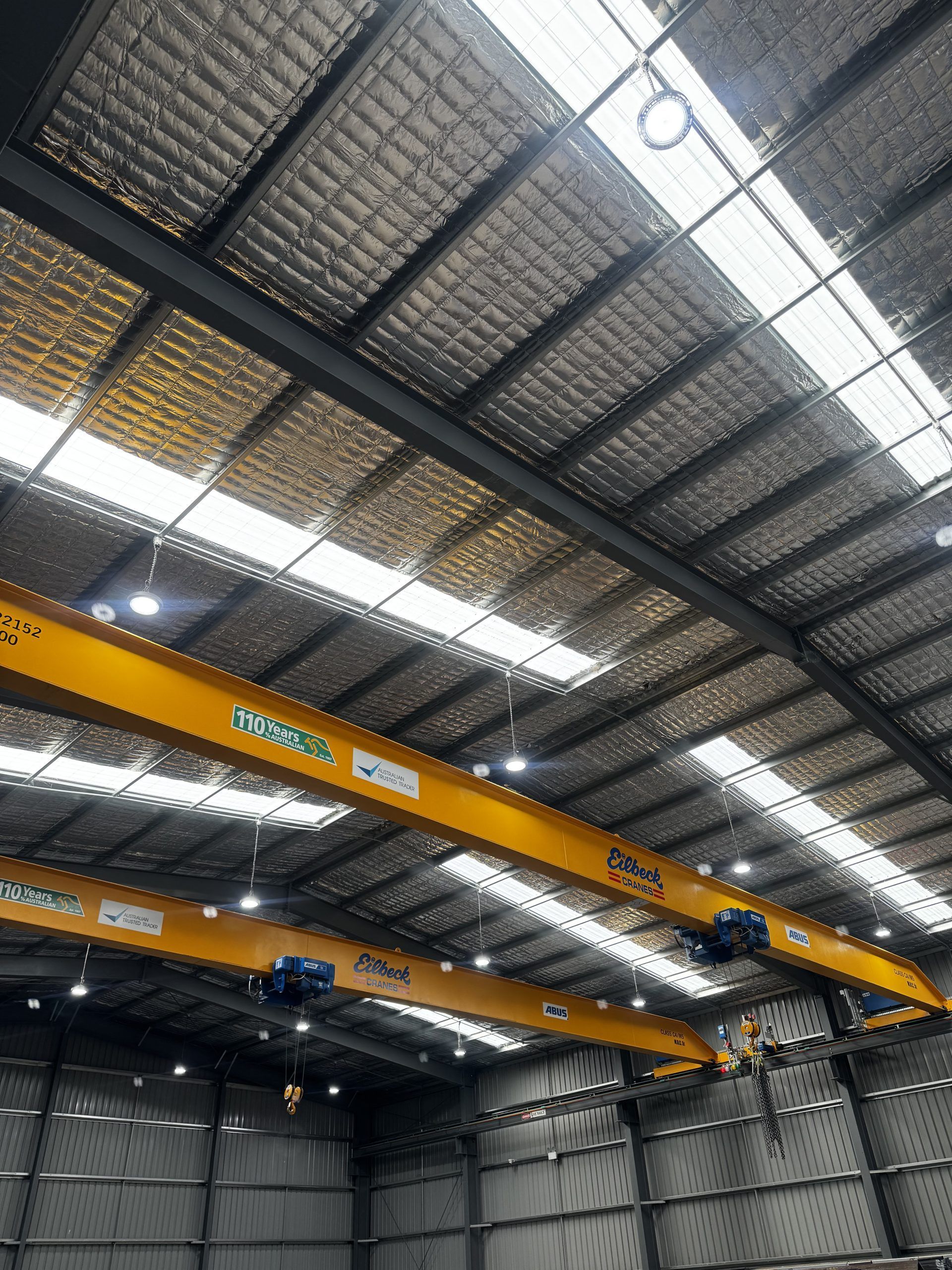 Inside a warehouse, overhead yellow gantry cranes span a high metal-roofed industrial space.
