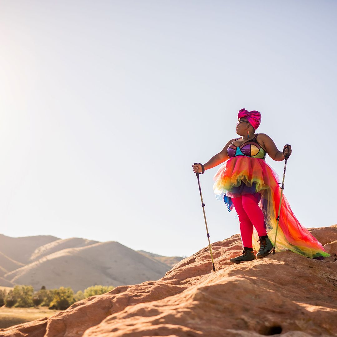 Woman wearing a rainbow tutu and hiking boots, holding trekking poles, and standing on the top of a mountain