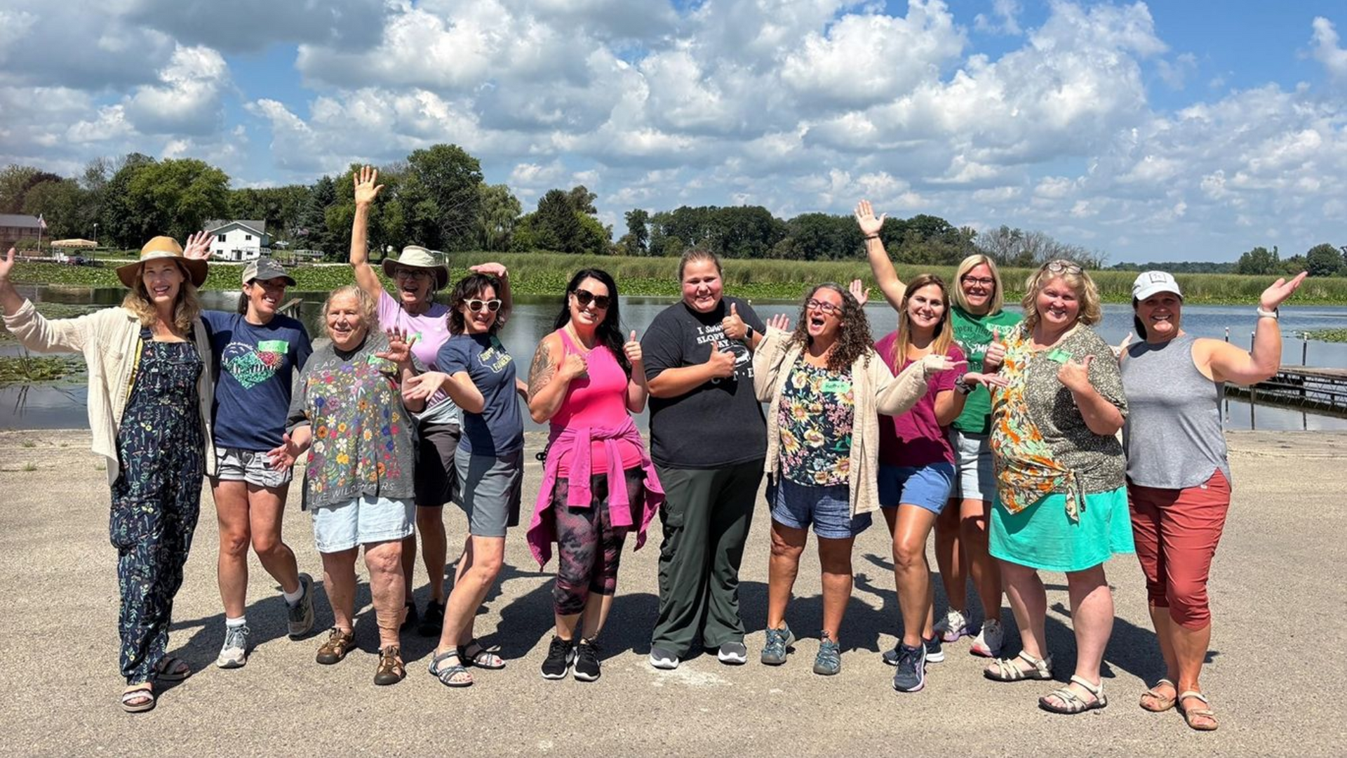 Group of people with arms raised and thumbs up on a sunny day, by a body of water.