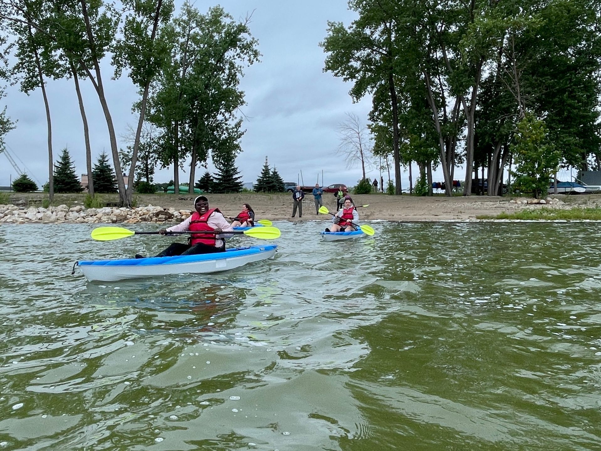 People kayaking on a lake near a sandy shore, overcast sky.