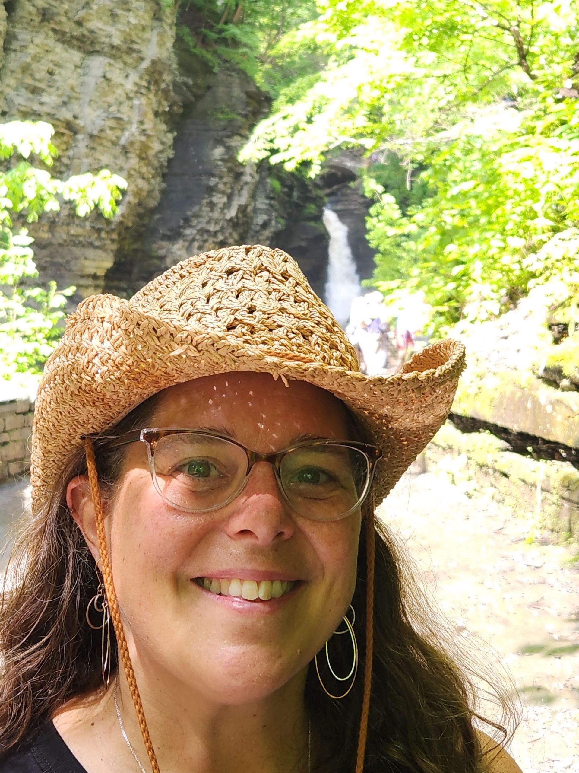 Woman in straw hat and glasses smiles, waterfall in background.