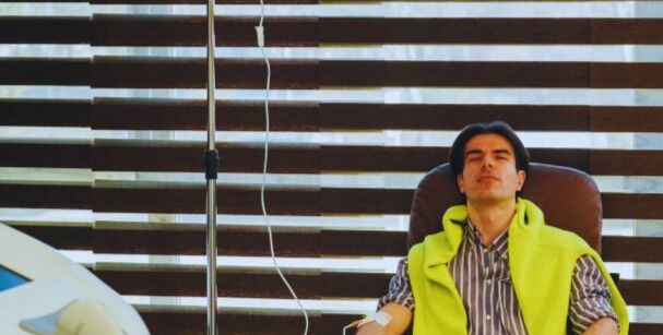 Person seated in a chair indoors, wearing a neon yellow vest and striped shirt, against horizontal blinds