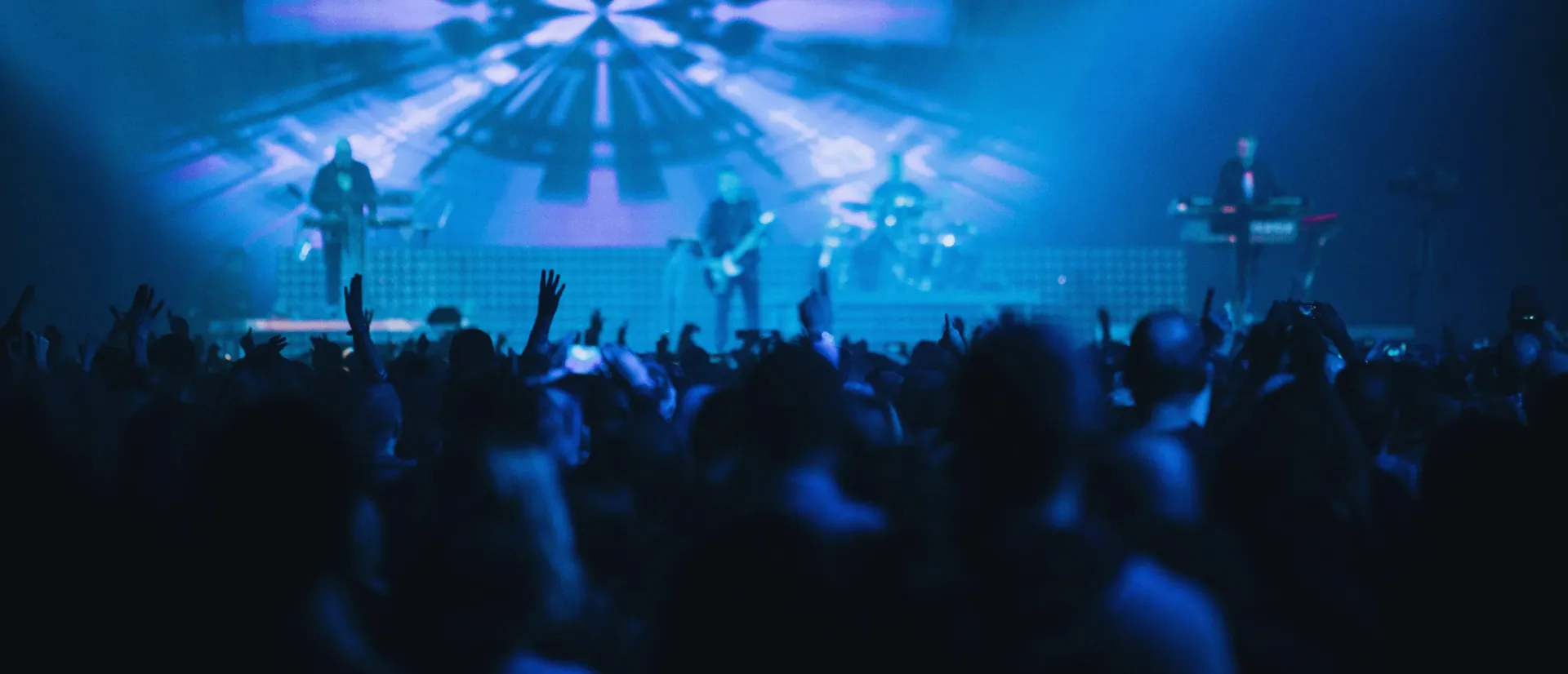 Blue-lit concert crowd with raised hands facing a performer on stage