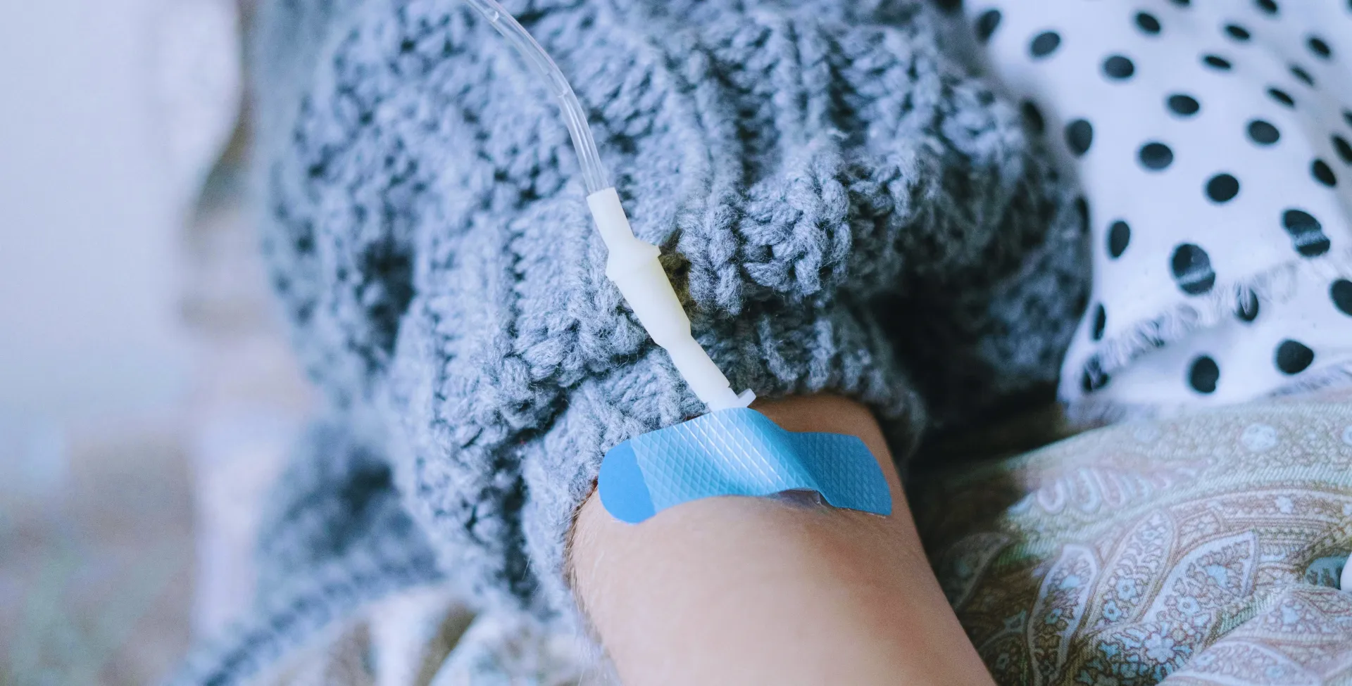 Person wearing a gray knit sweater has a blue bandage on an arm, with a polka-dot pillow in the background