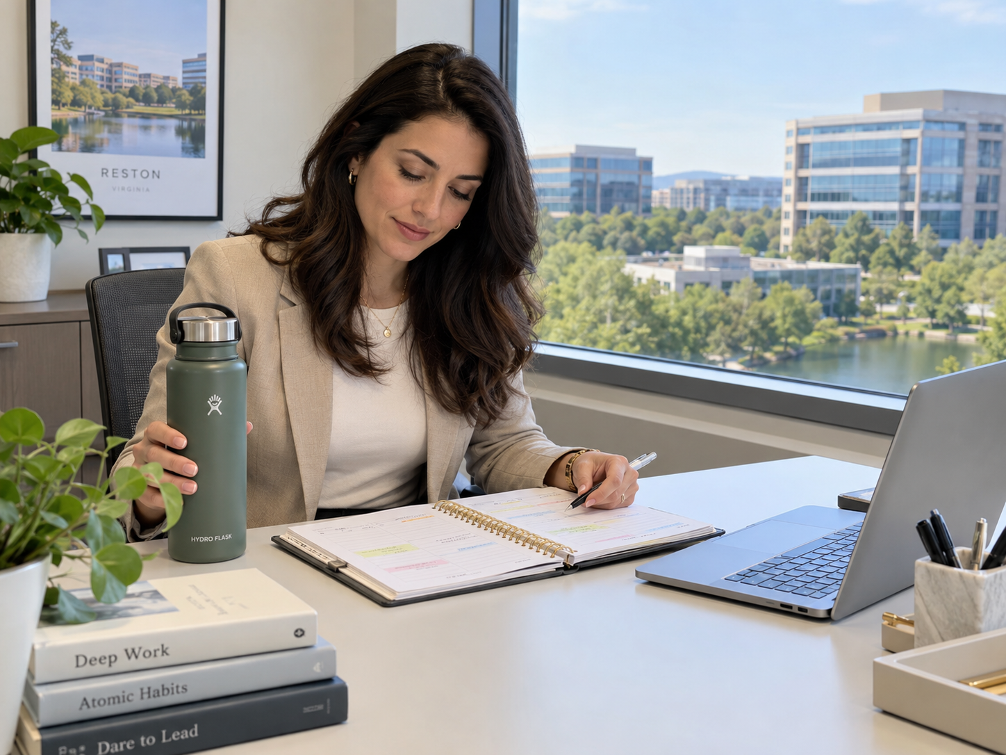 Woman writing in a notebook at a desk by a window, with a laptop and water bottle in a bright office.