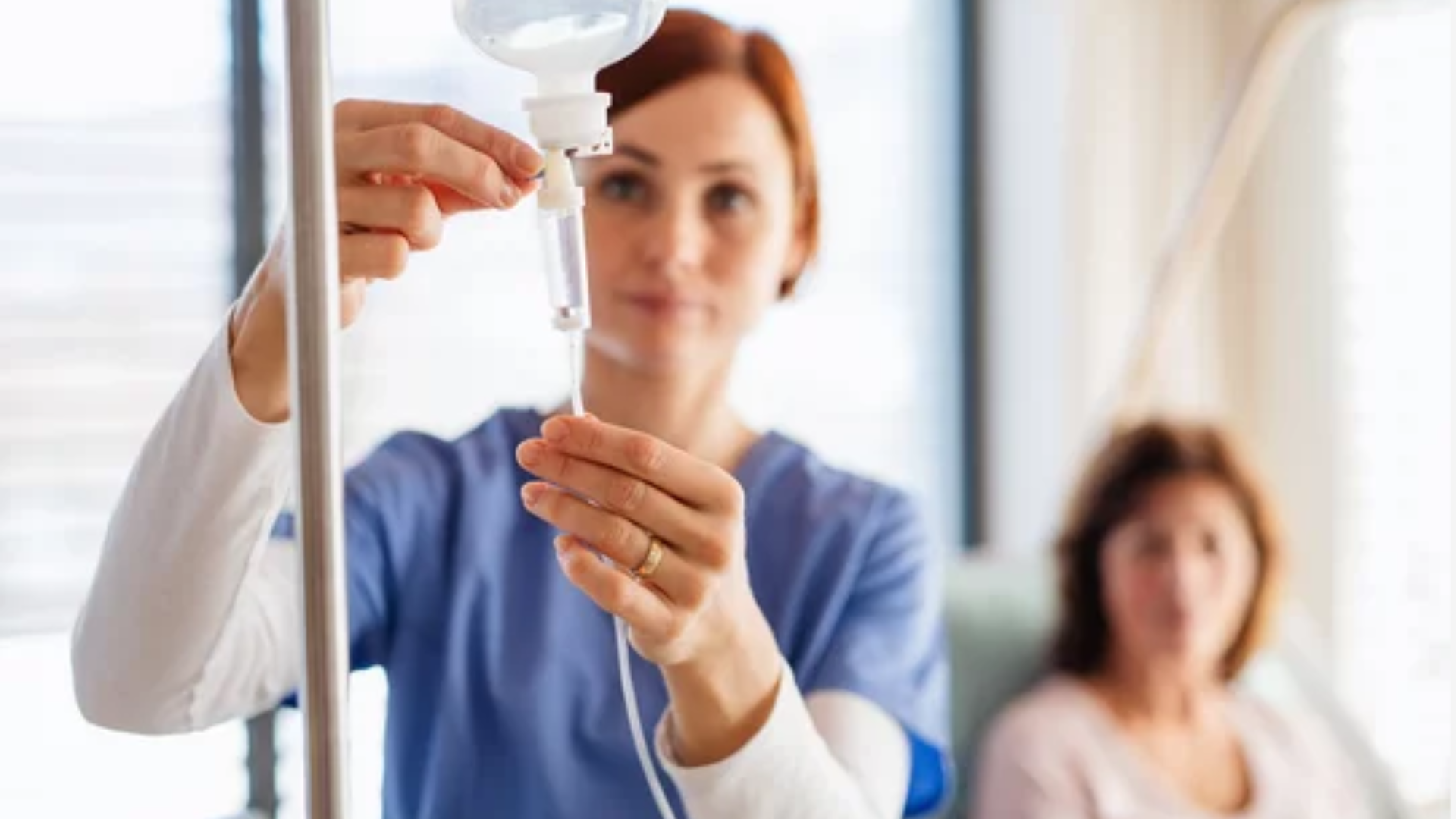 Nurse filling an IV drip in a hospital room, with a patient sitting blurred in the background
