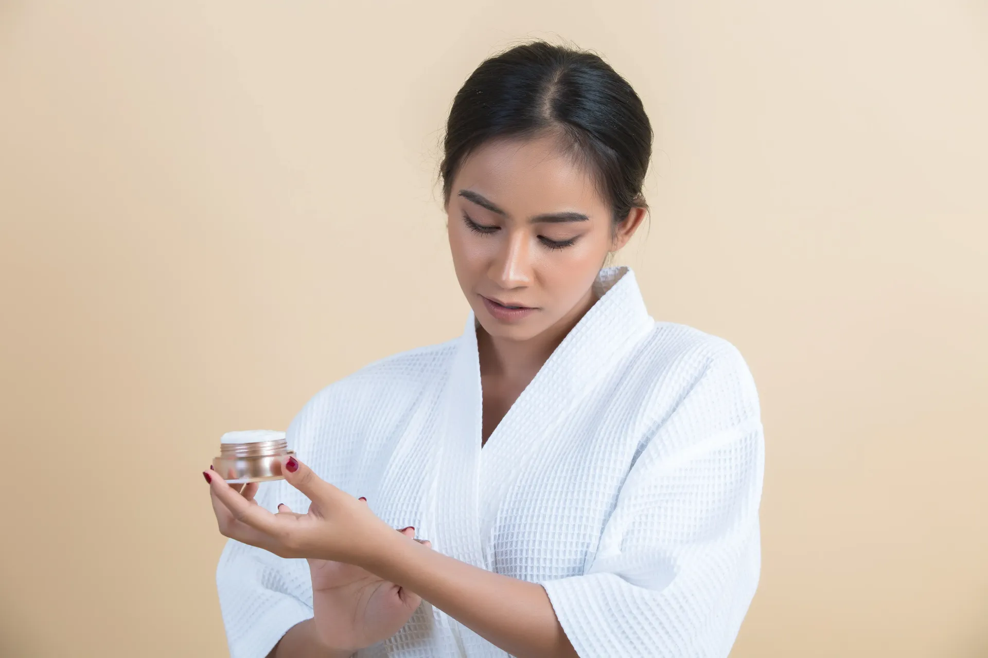 Woman in white robe examining cream jar against a beige background.