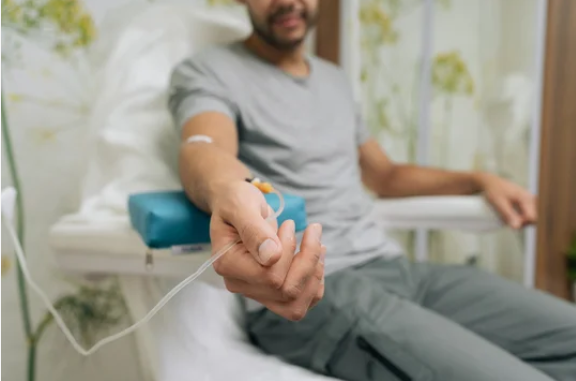 Person receiving IV fluids in a medical setting, seated with arm extended, white tube attached.
