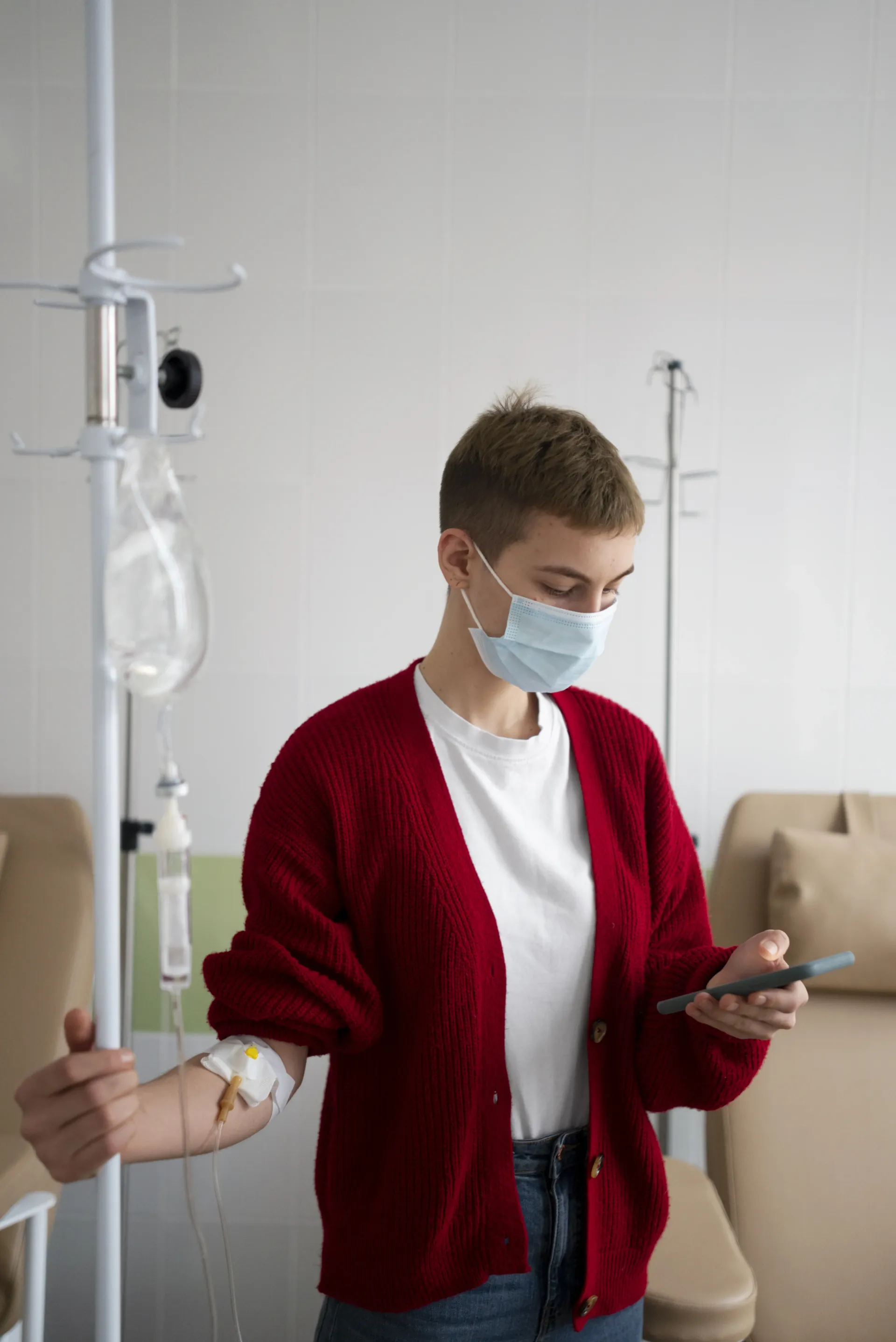 Masked medical worker holding a phone beside an IV stand in a clinic room