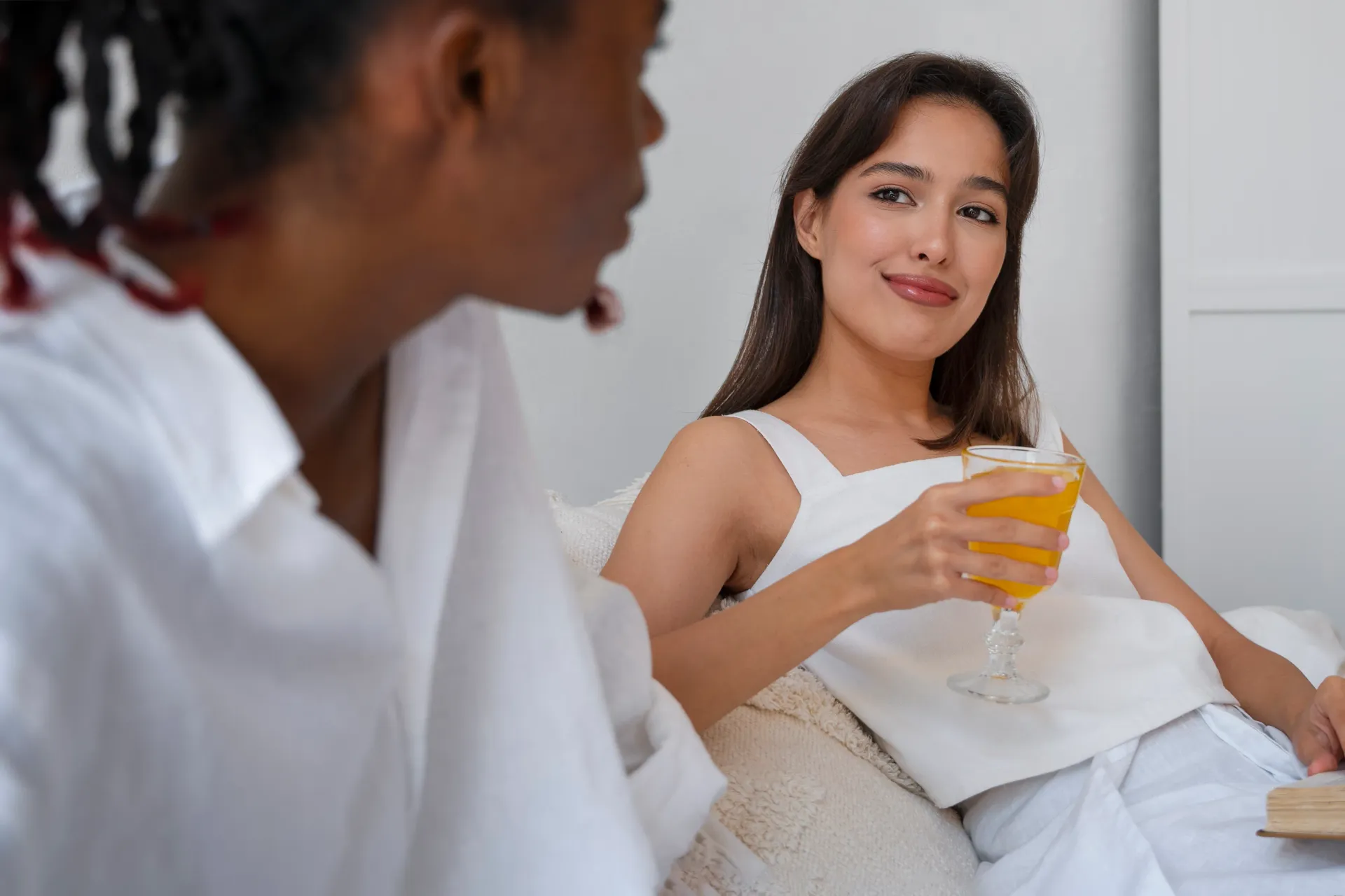 Woman reclining on a couch, holding a glass of orange drink while speaking with another person in the foreground