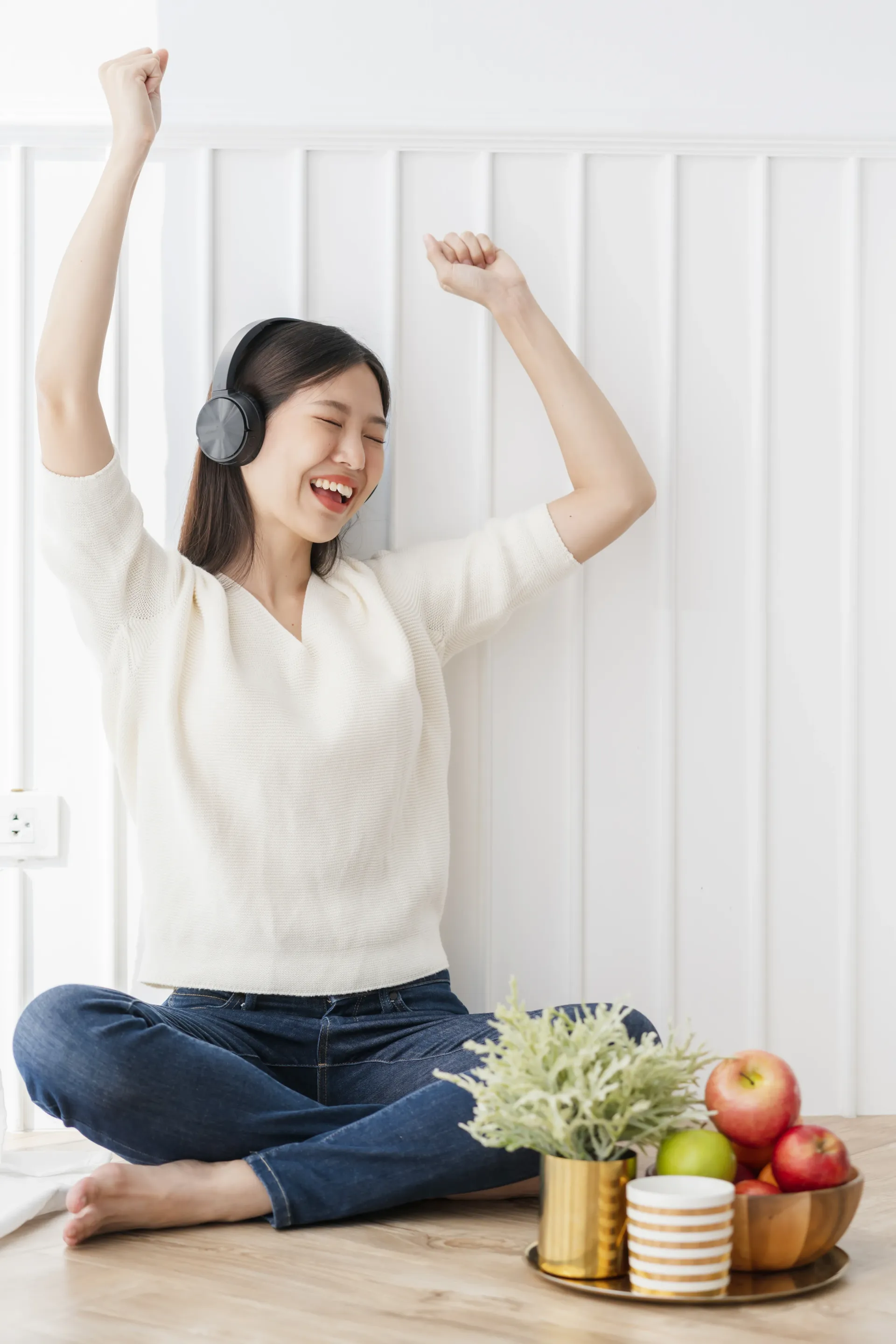 Person dancing on the floor beside fruit and a potted plant, wearing headphones and a white sweater