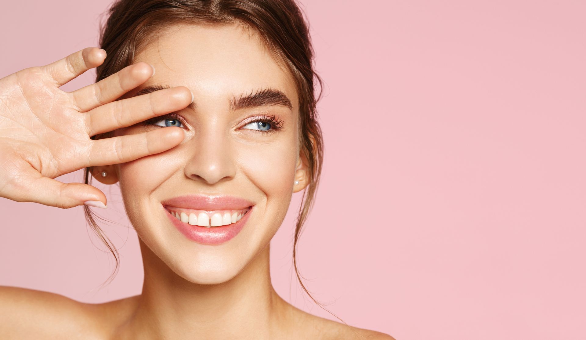 Smiling woman posing against a pink background while covering one eye with her hand