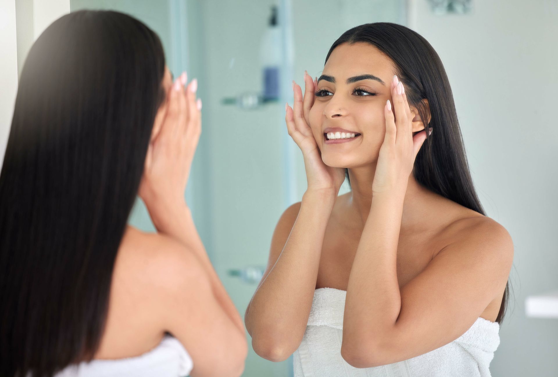 Woman smiling at bathroom mirror while applying skincare with both hands on her cheeks