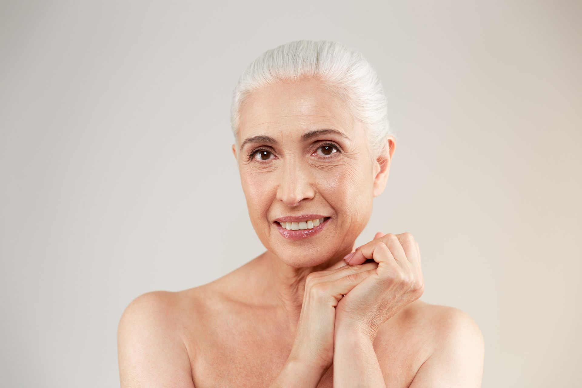 Woman with short white hair smiles, hands clasped under chin, bare shoulders, soft lighting.