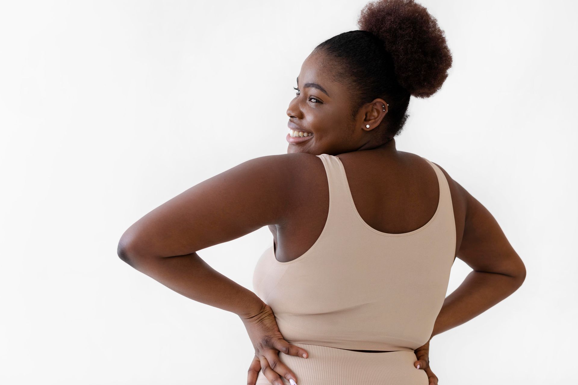 Woman with brown skin smiles, poses with hands on hips, wearing beige tank top and pants.