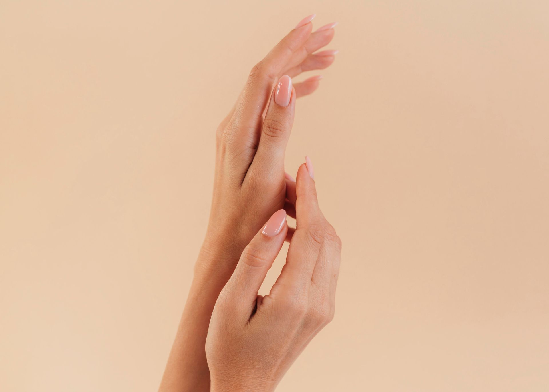 Hands with pink nail polish against a beige background, one hand gently touching the other.