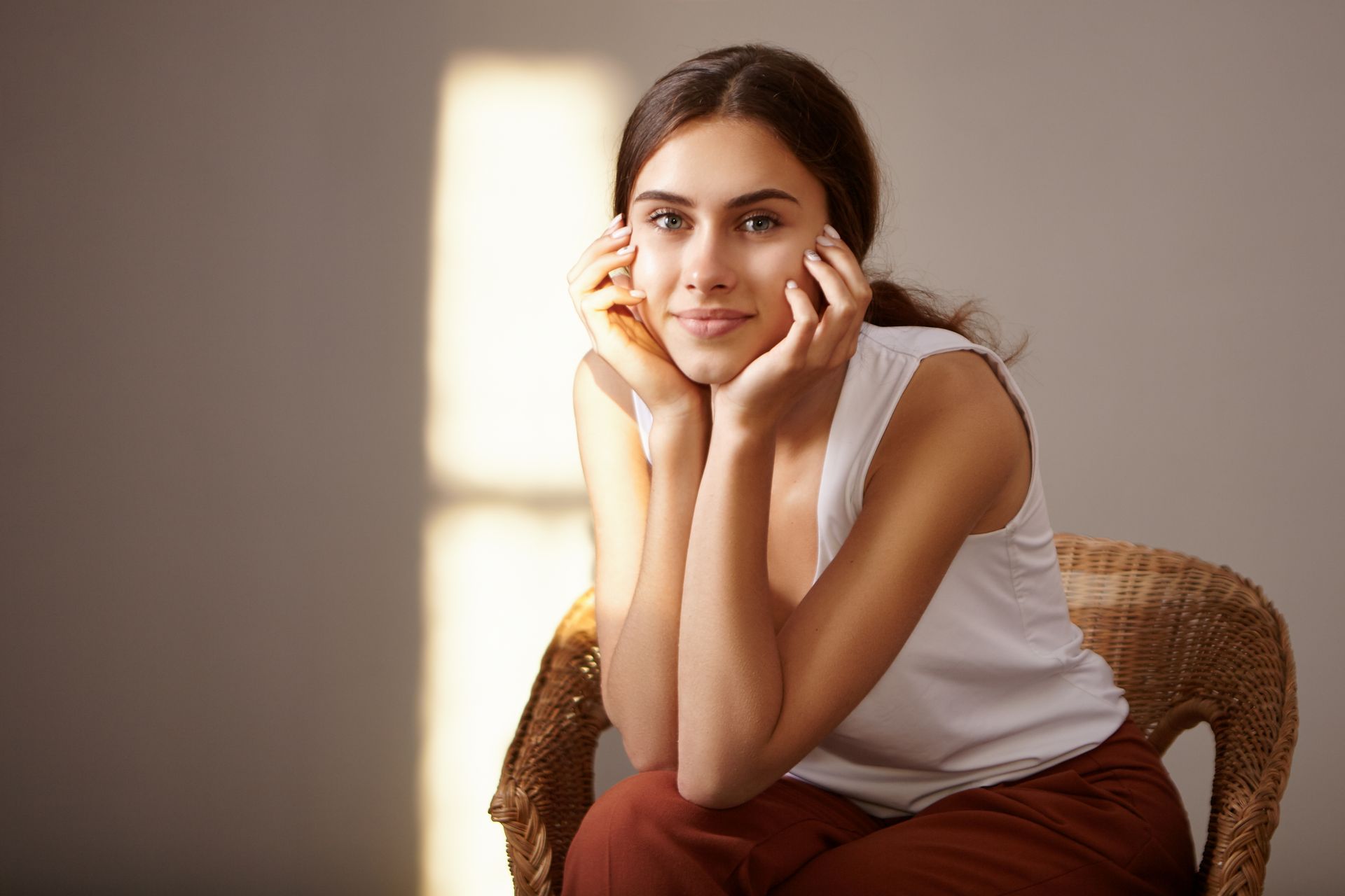 Woman sitting in a wicker chair, resting her face in her hands by a sunlit wall.