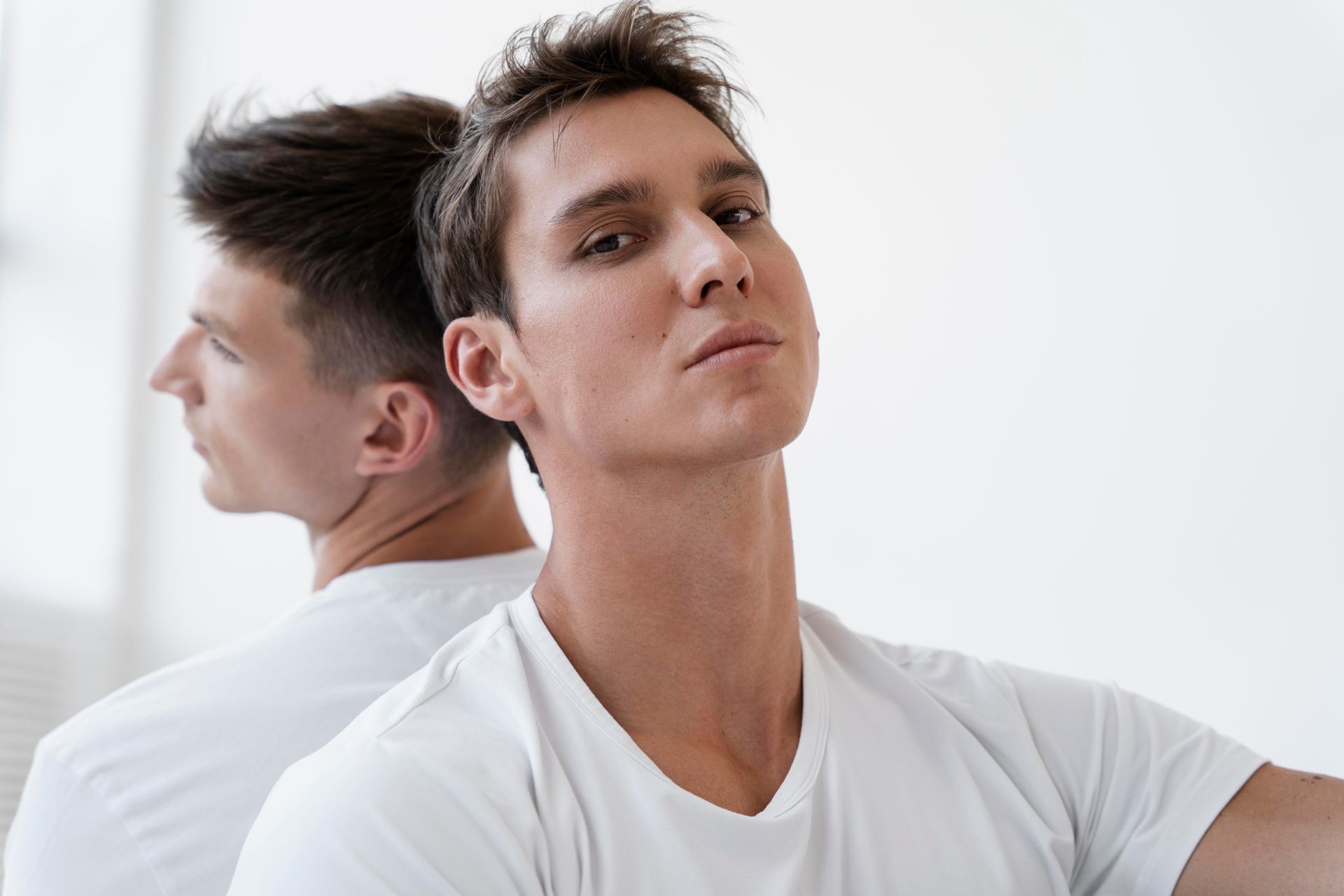 A man in a white t-shirt looks at the viewer, another man behind him looking away; white background.