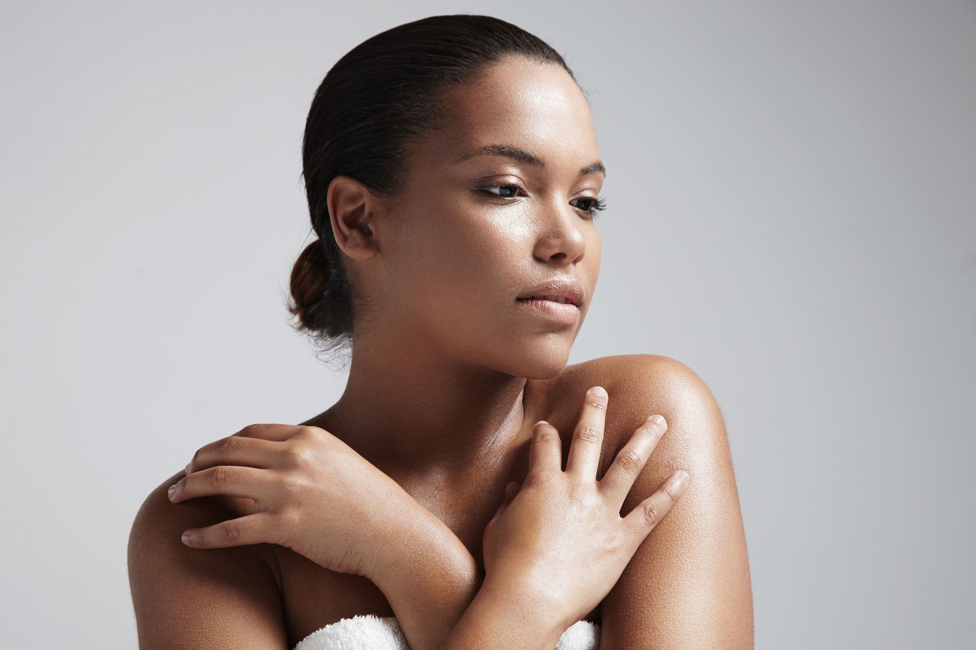 Woman with glowing skin, hands crossed on shoulders, looking away, neutral background.